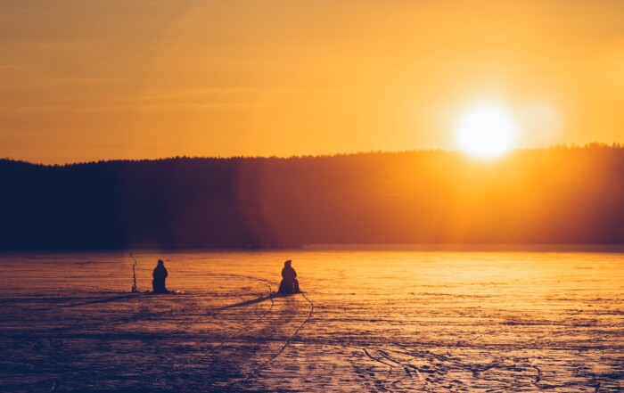 Silhouette Of Two Persons Sitting While Snow Fishing On An Iced Covered Body Of Water At Dawn
