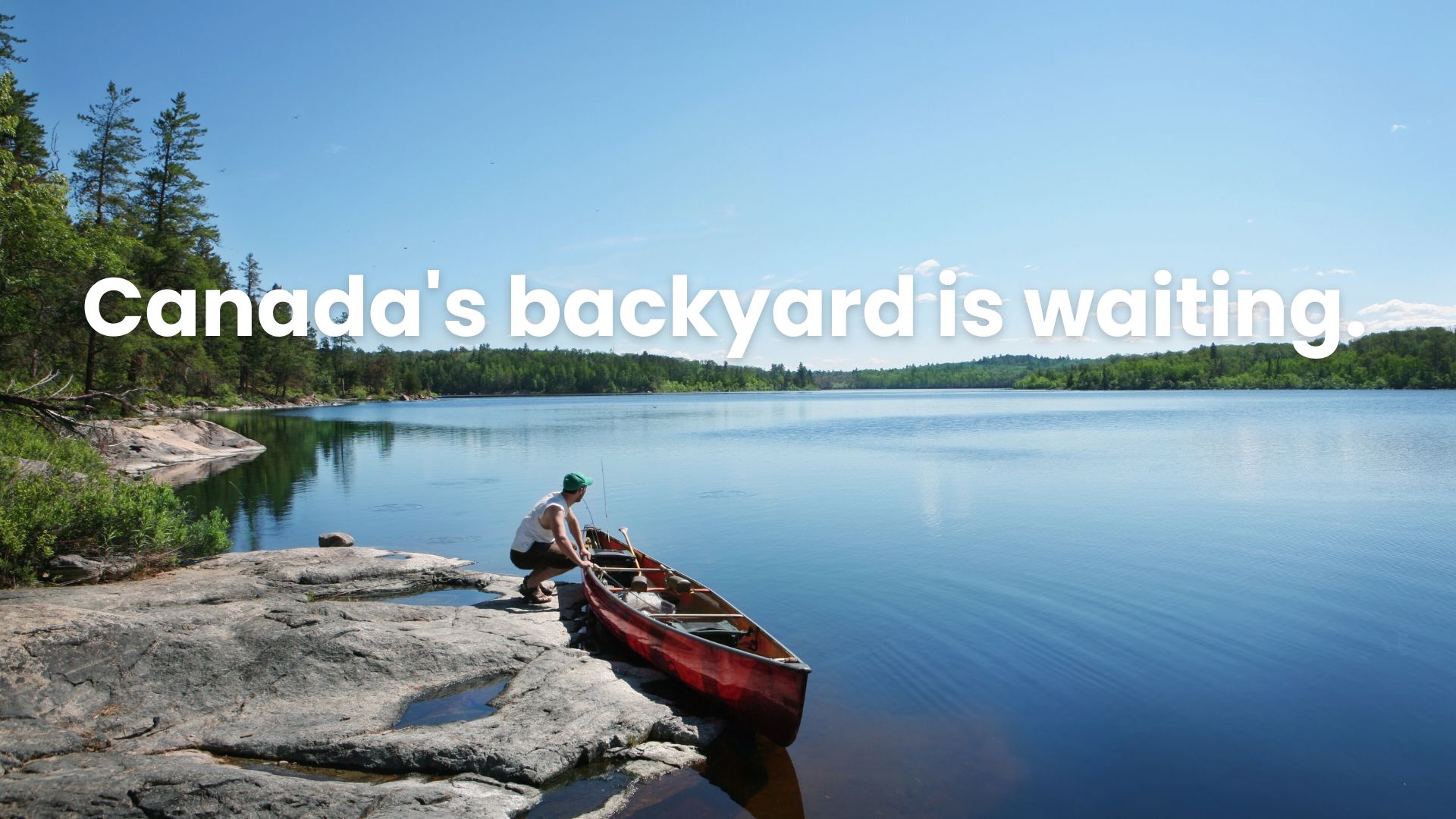 A person stands at the edge of the Canadian shield by a canoe, looking out onto a lake.