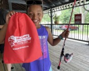 A smiling girl holds a bag with a Kids and Cops logo in one hand, and a fishing rod in the other hand.