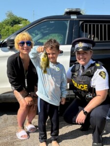 A little boy stands between two adults, one of whom is a police officer, holding up a big fish